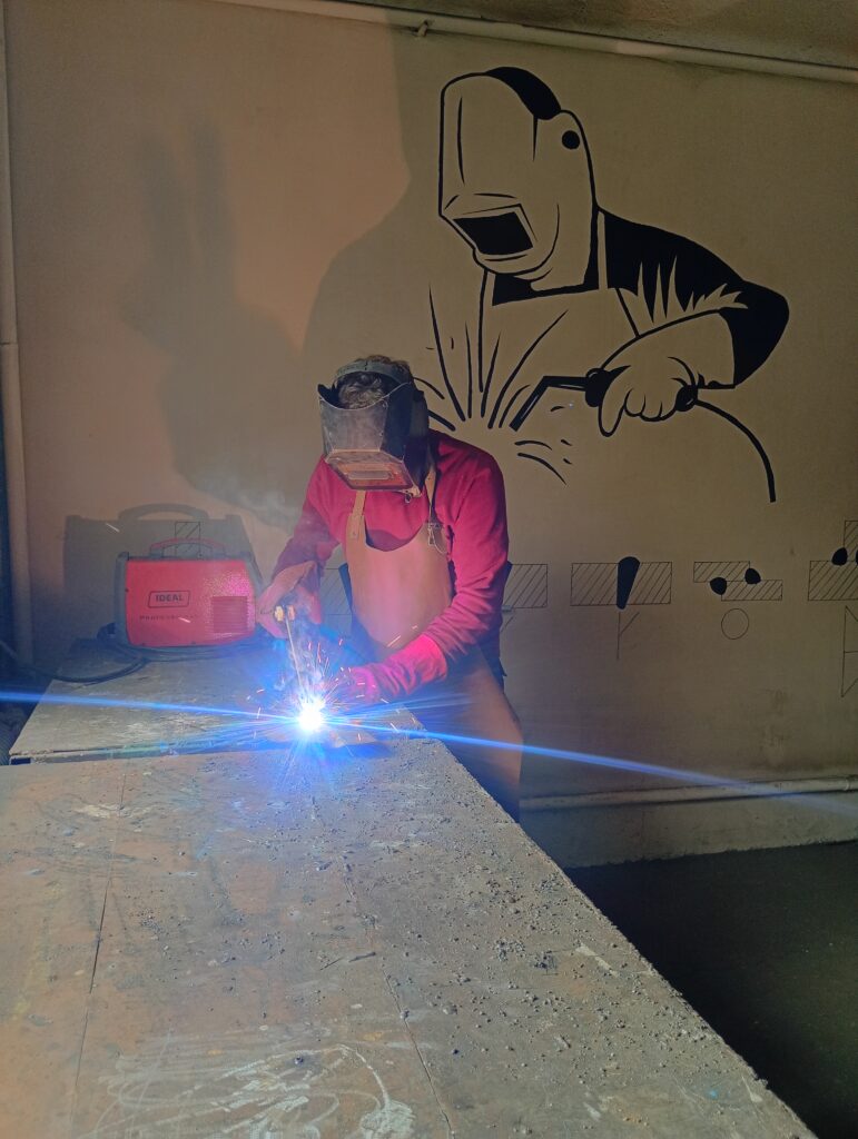 Spawacz w maskach ochronnych wykonuje spawanie, a na ścianie za nim znajduje się malowidło przedstawiające sylwetkę spawacza.