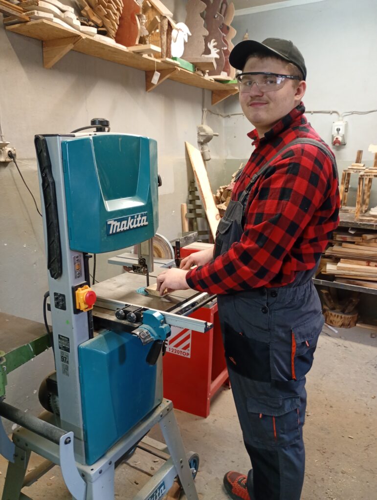 Mężczyzna w okularach ochronnych i roboczym ubraniu pracuje przy pile taśmowej w warsztacie stolarskim.