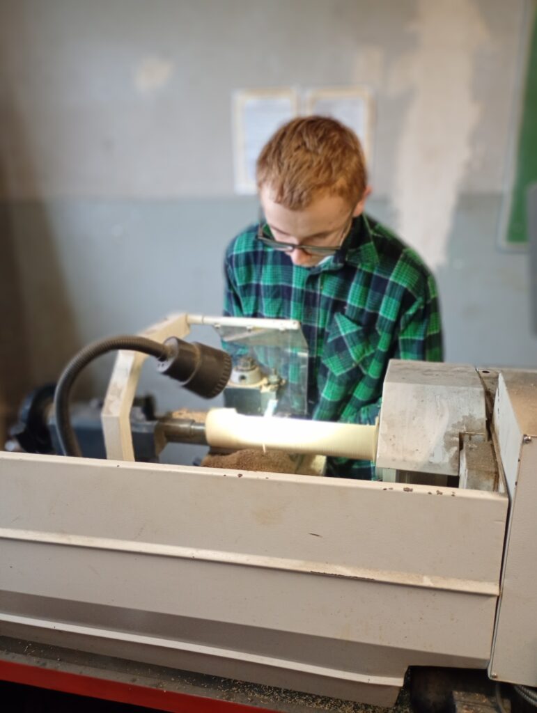 Chłopiec w zielonej koszuli pracuje na tokarce, obrabiając drewniany wałek.