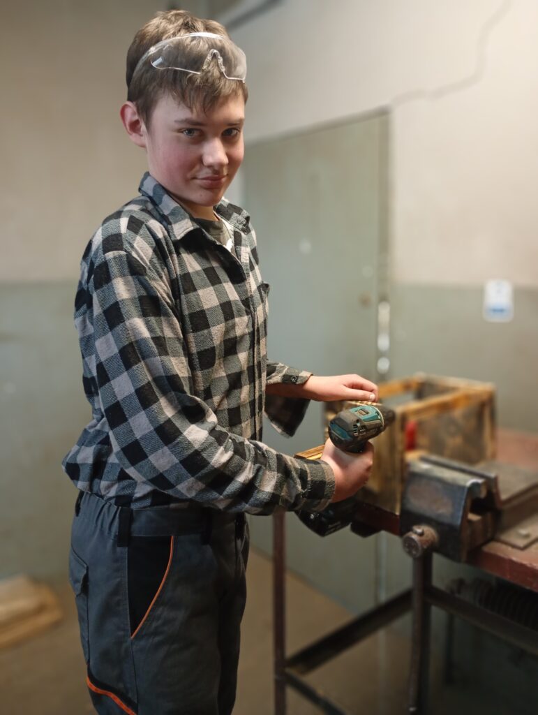 Chłopak w okularach ochronnych trzyma wiertarkę i pracuje przy drewnianym elemencie zamocowanym w imadle.