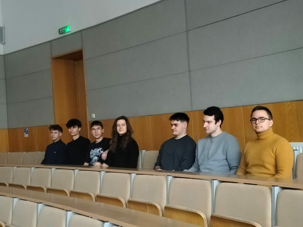 Siedem osób siedzi w rzędzie ławek w sali wykładowej.