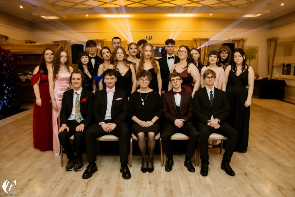 Grupa młodych osób ubranych w eleganckie suknie i garnitury pozująca do zdjęcia w sali balowej.