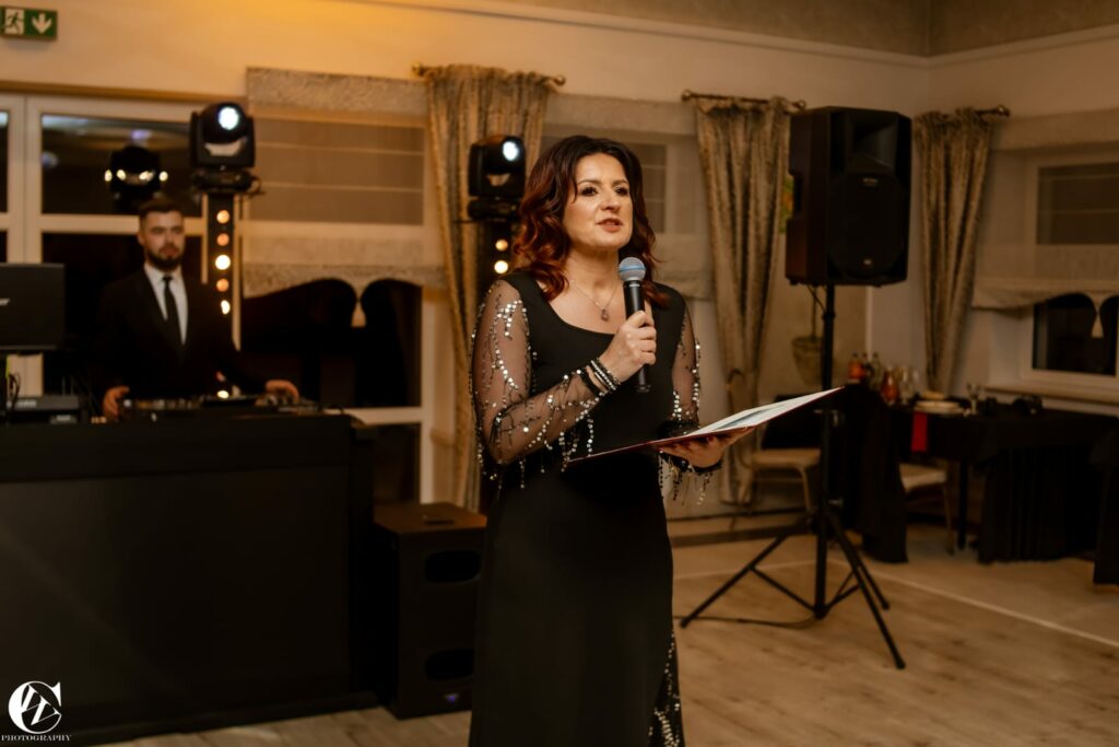 Kobieta w eleganckiej sukni przemawia do mikrofonu, trzymając w ręku otwartą teczkę, a w tle znajduje się mężczyzna przy konsoli DJ-a.