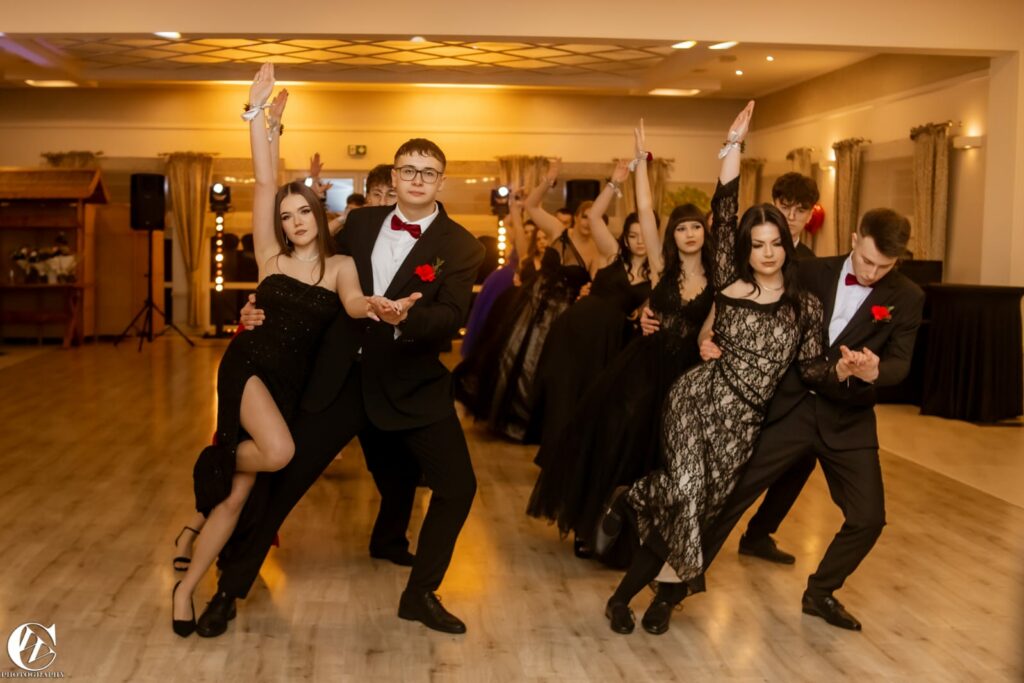 Młode pary w eleganckich strojach wykonują wspólnie taniec w sali balowej o ciepłym oświetleniu.