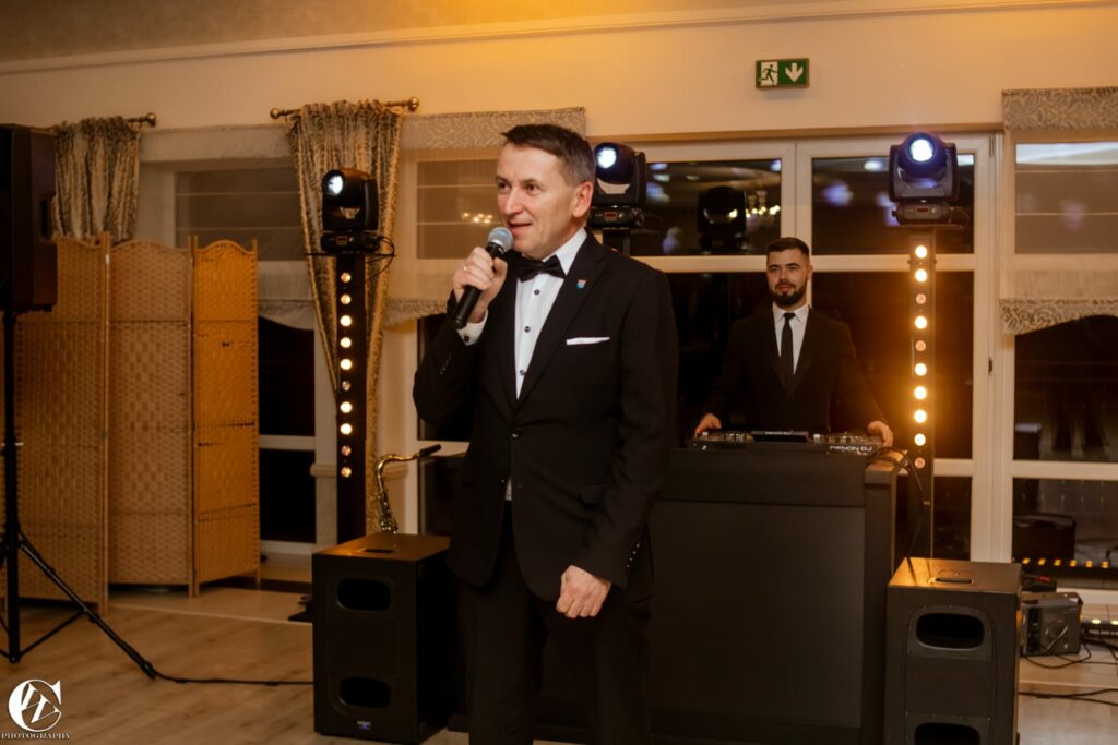 Mężczyzna w eleganckim garniturze przemawia do mikrofonu podczas uroczystości, a za nim stoi DJ obsługujący sprzęt muzyczny.