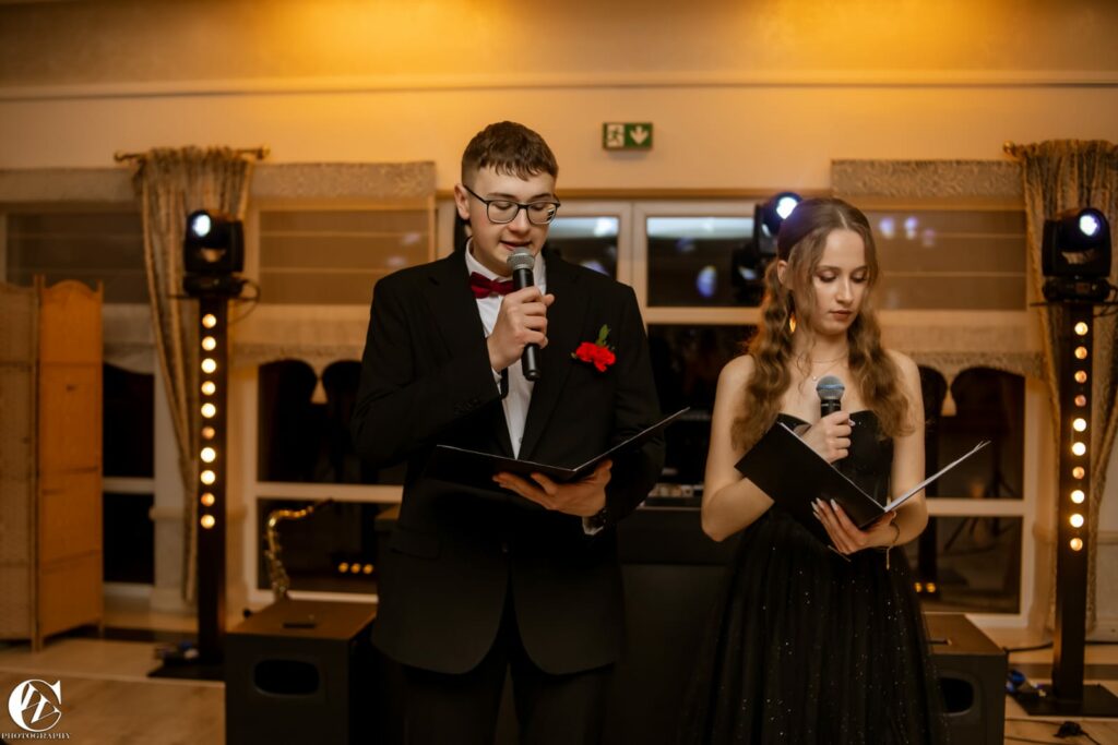 Dwójka młodych ludzi w eleganckich strojach prowadzi wydarzenie, trzymając mikrofony i kartki z tekstem.