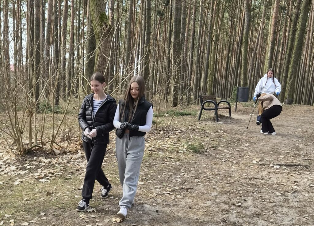 Cztery młode osoby sprzątają las, zbierając śmieci do worków, na tle drzew i ławki.