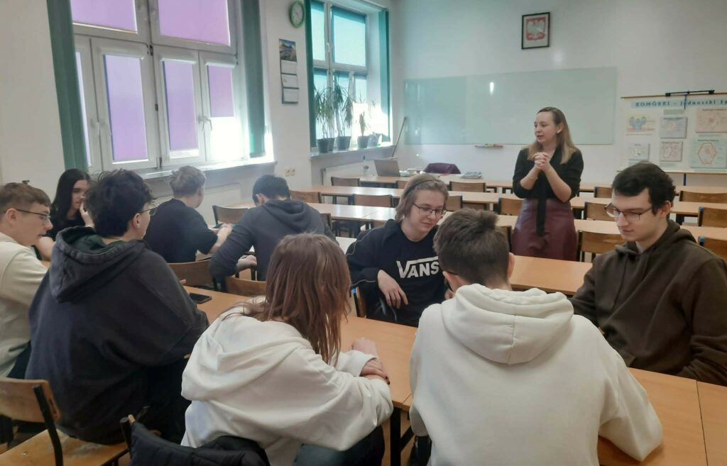 Grupa uczniów siedzi przy stołach w klasie, a nauczycielka stoi przed tablicą, prowadząc lekcję.