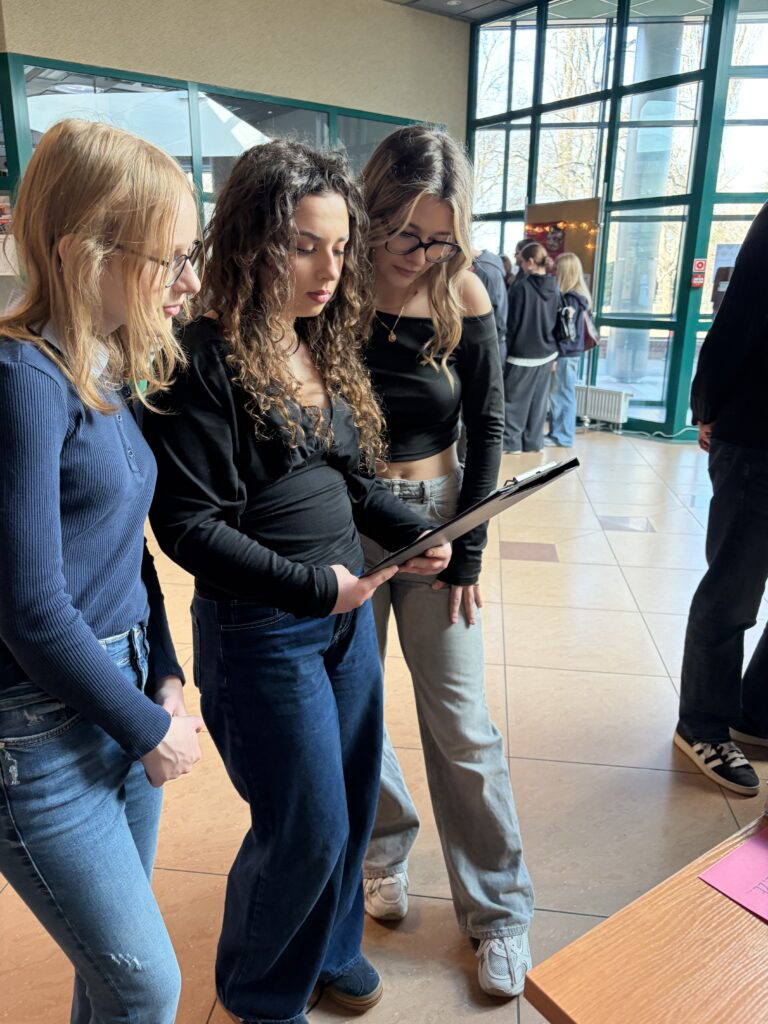 Trzy młode kobiety stoją razem, jedna z nich trzyma teczkę i razem coś czytają w jasnym pomieszczeniu z dużymi oknami.