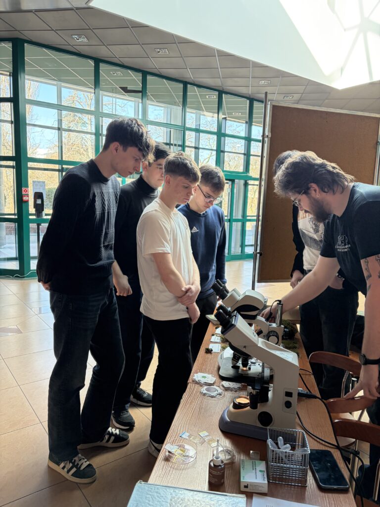 Grupa młodych mężczyzn przygląda się mikroskopom na stole, podczas gdy jedna osoba z prawej strony tłumaczy lub pokazuje coś związanego z ich użyciem.