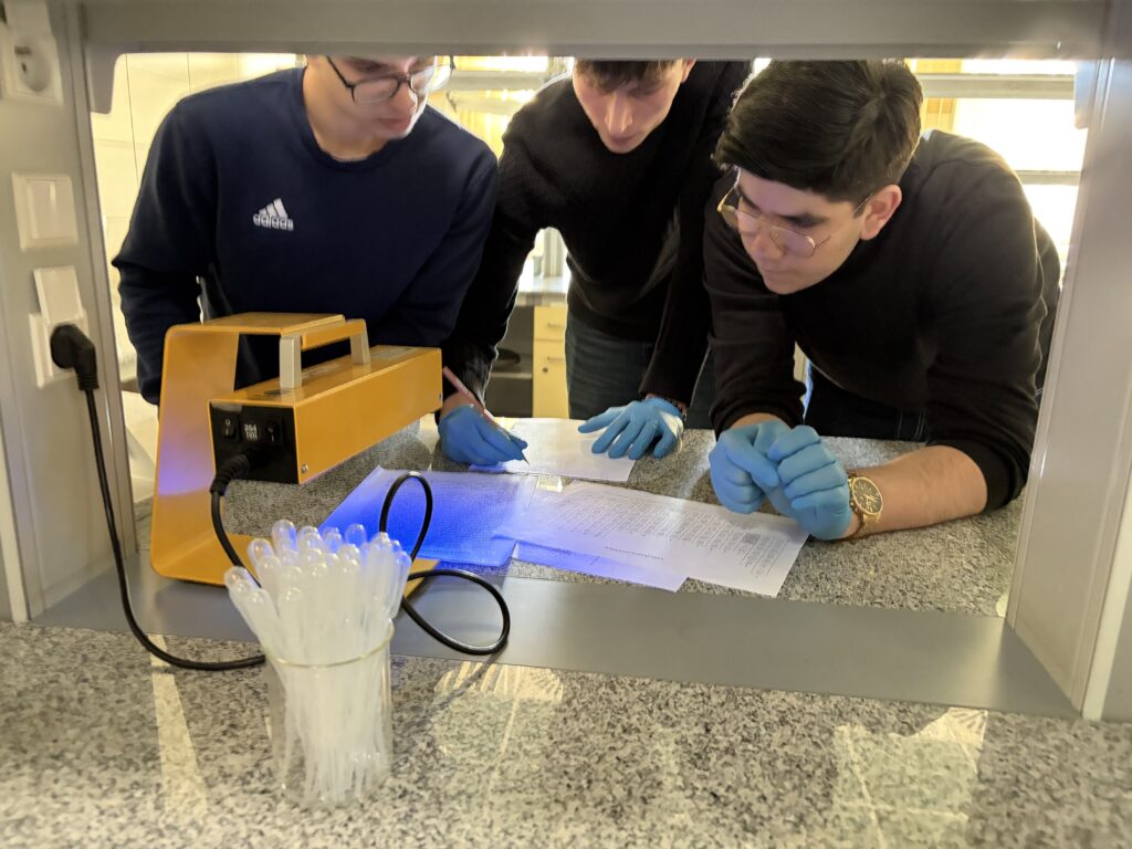 Trzech młodych mężczyzn w rękawiczkach niebieskich analizuje i notuje dane na kartach przy stole laboratoryjnym z urządzeniem emitującym niebieskie światło.