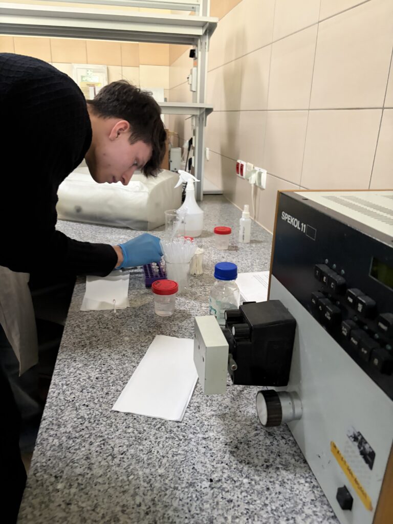 Młody mężczyzna w laboratorium wykonuje precyzyjne czynności z pipetą, obok stoi sprzęt laboratoryjny i różne pojemniki na blacie.