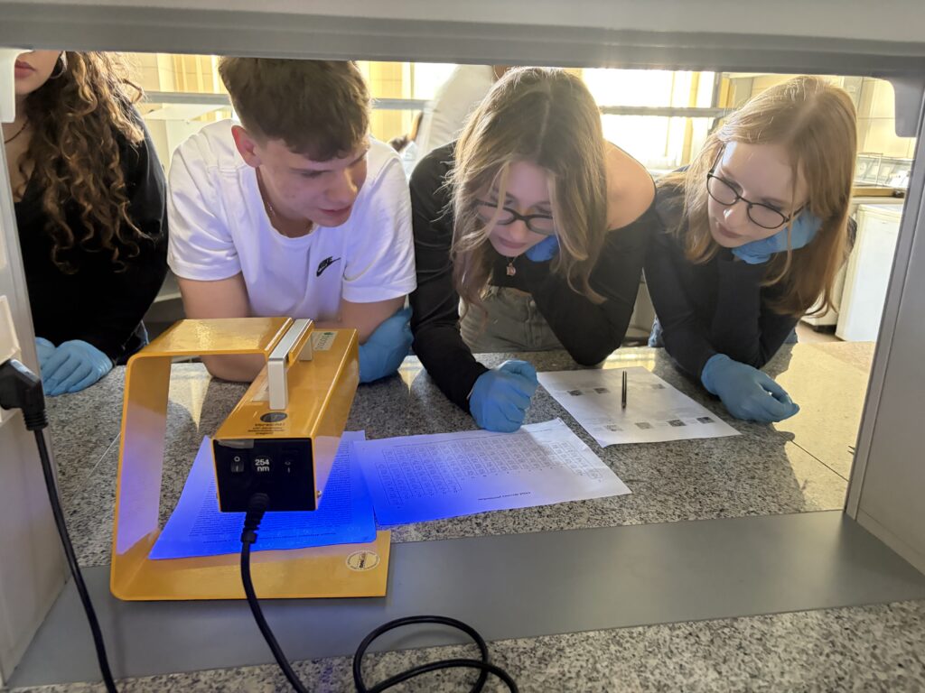 Grupa młodych ludzi w rękawiczkach ochronnych z uwagą analizuje kartę okresową pierwiastków przy świetle lampy UV na blacie laboratoryjnym.