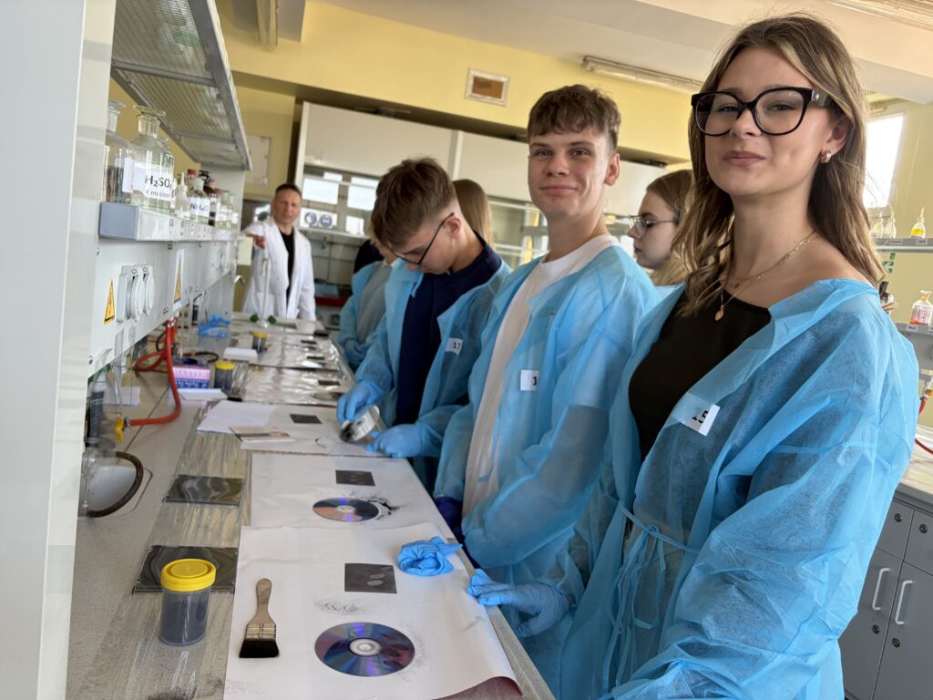 Grupa młodych osób w niebieskich fartuchach ochronnych i rękawiczkach wykonuje ćwiczenia z zakresu kryminalistyki w laboratorium, rozkładając odciski palców na płytach CD i innych powierzchniach.