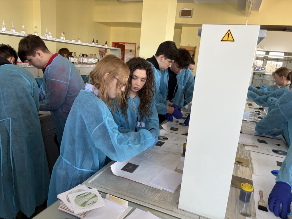 Grupa młodych osób ubranych w niebieskie fartuchy ochronne wykonuje eksperymenty w laboratorium chemicznym.