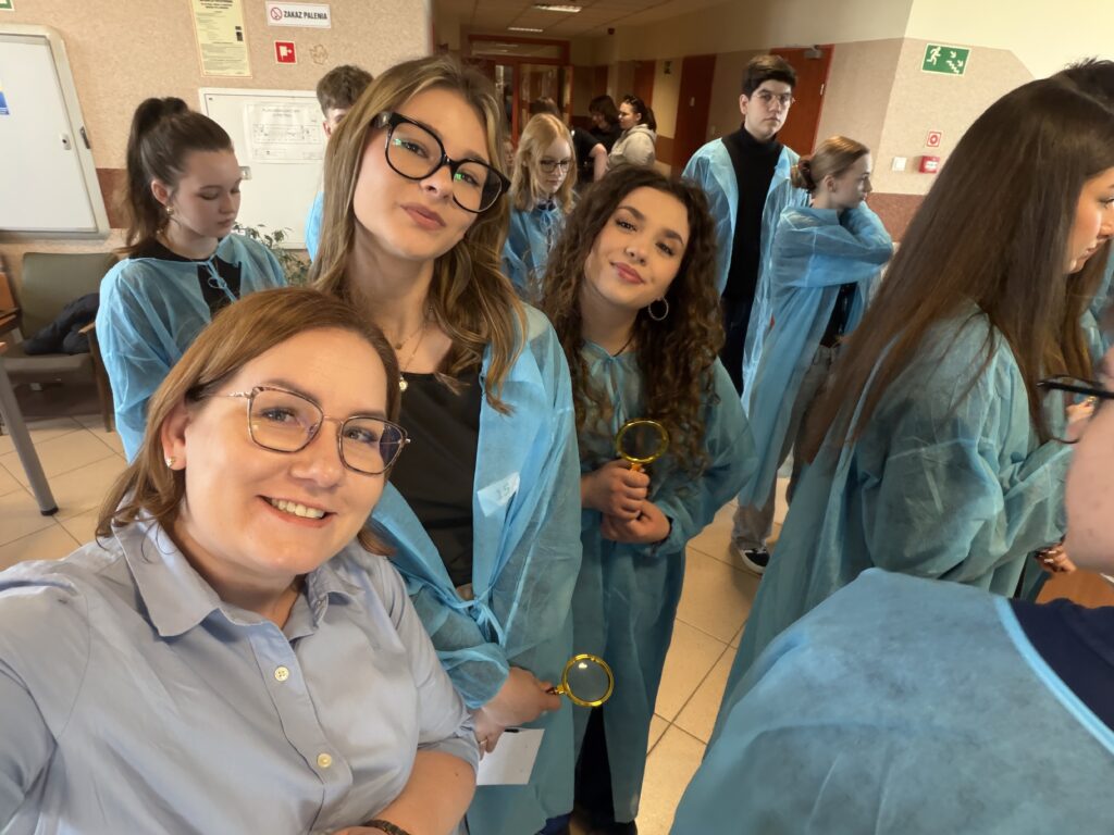 Grupa młodych osób w niebieskich fartuchach ochronnych, niektórzy trzymają lupy, stoją w pomieszczeniu o szpitalnym lub szkolnym charakterze.