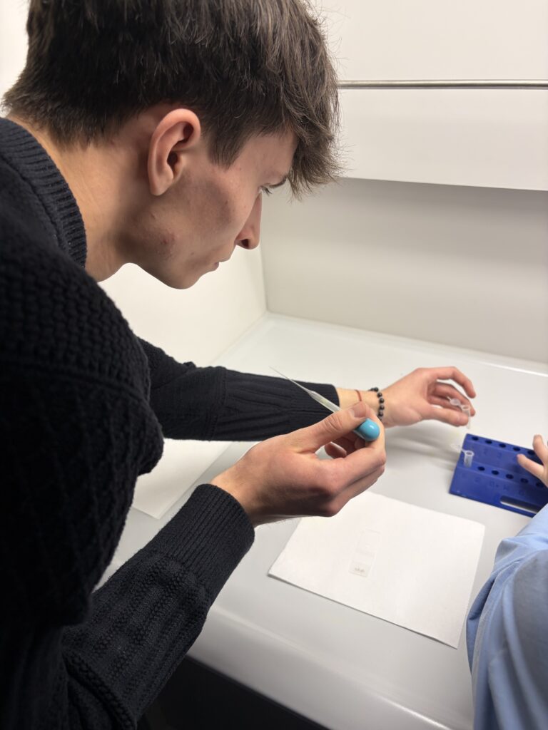 Młody mężczyzna w czarnym swetrze pracuje z pipetą i probówkami w laboratorium.