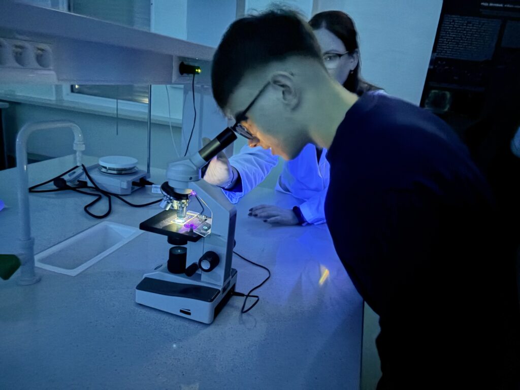 Młody człowiek obserwuje próbkę pod mikroskopem w ciemnym laboratorium, a obok stoi osoba w białym fartuchu.