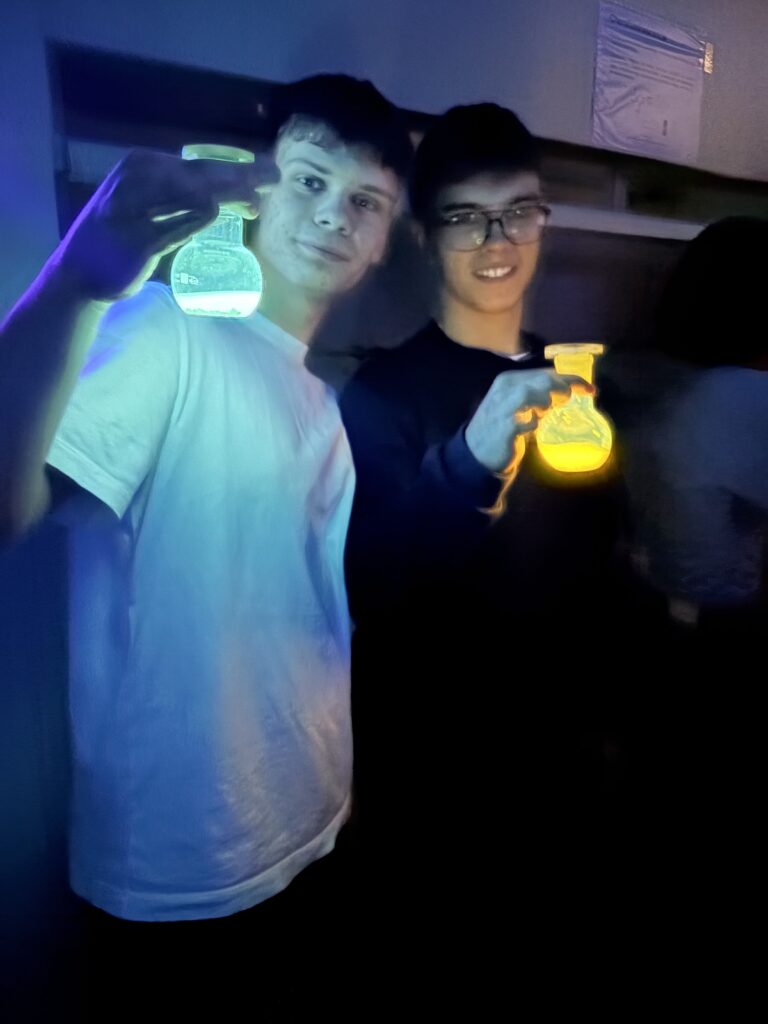 Dwóch młodych mężczyzn trzyma w rękach świecące w ciemności kolby laboratoryjne – jedna zielona, druga żółta.