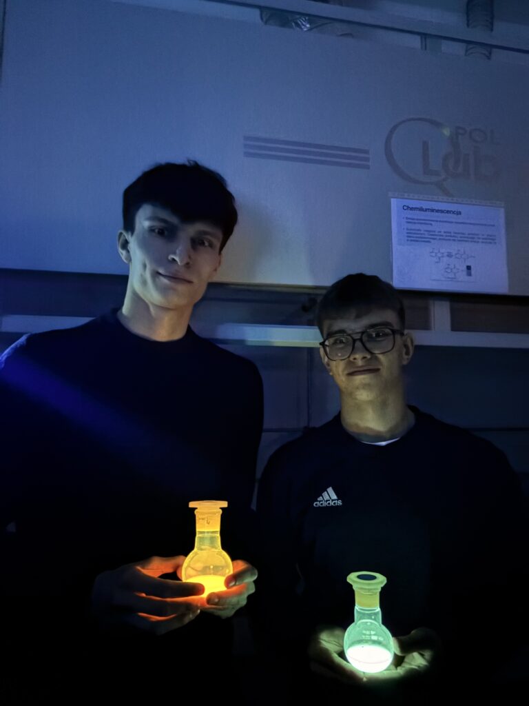Dwaj młodzi mężczyźni trzymają w ciemności świecące kolbówki z chemiluminescencyjnymi roztworami – jedna świeci na pomarańczowo, druga na zielono.