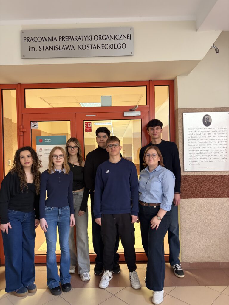 Grupa młodych osób stoi przed drzwiami pracowni preparatyki organicznej im. Stanisława Kostaneckiego w budynku uczelni.