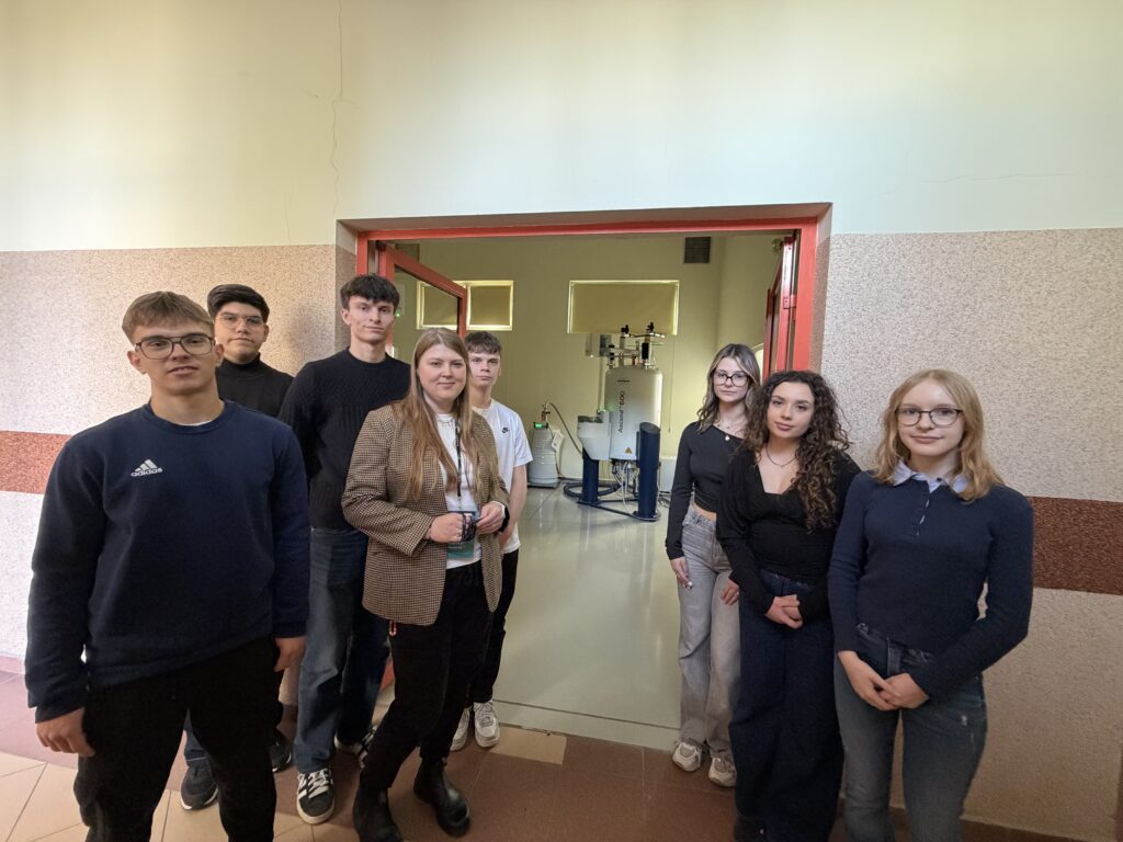 Grupa młodych ludzi stoi w korytarzu przed otwartymi drzwiami do pracowni z dużym, zaawansowanym urządzeniem naukowym w tle.