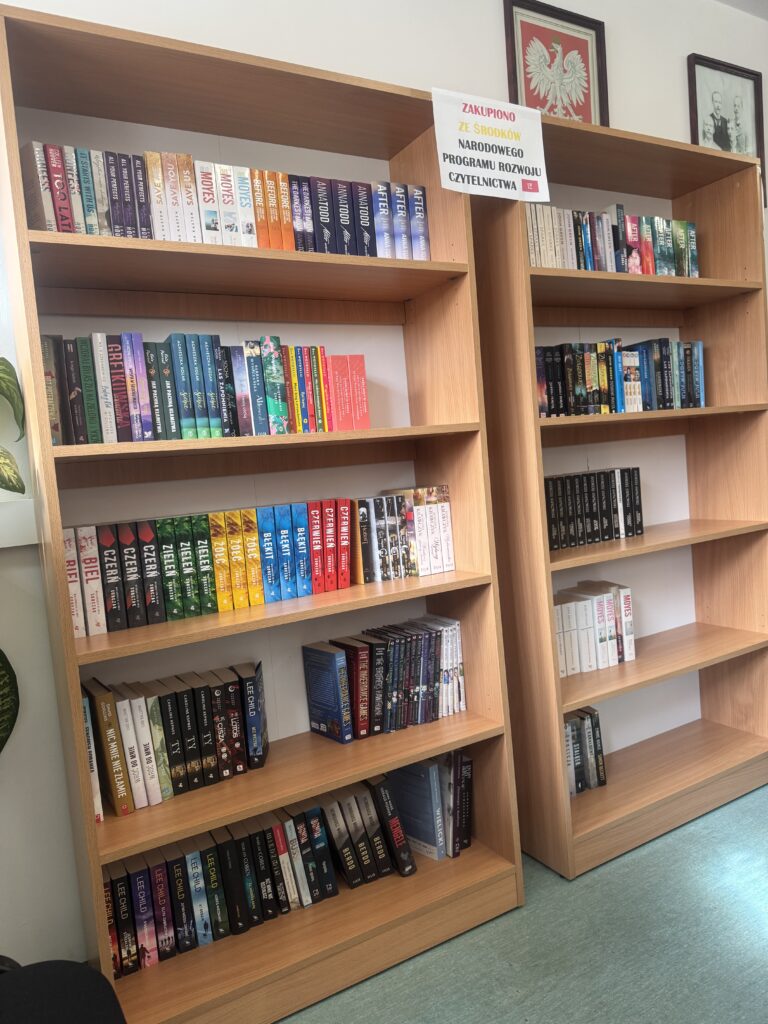 Drewniane regały z książkami w różnorodnych kolorach, z tabliczką informującą o zakupie ze środków Narodowego Programu Rozwoju Czytelnictwa.