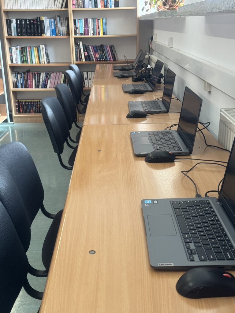 Rząd laptopów z myszkami na drewnianym stole ustawionym naprzeciwko czarnych krzeseł w pomieszczeniu z regałami pełnymi książek.