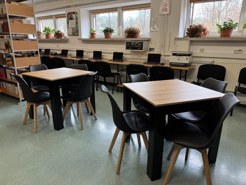 W czytelni znajdują się stoły do pracy z czterema krzesłami oraz rząd laptopów na długim stole przy oknie, a na parapetach stoją doniczki z roślinami.