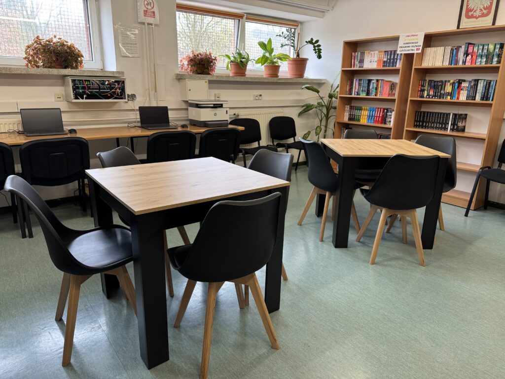 Sala z dwoma stołami i krzesłami, regałem z książkami, laptopami na długim biurku oraz roślinami na parapetach.