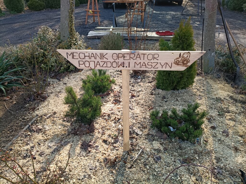 Drewniana tabliczka z napisem "MECHANIK OPERATOR POJAZDÓW I MASZYN" oraz rysunkiem maszyny rolniczej, umieszczona w ogrodzie.