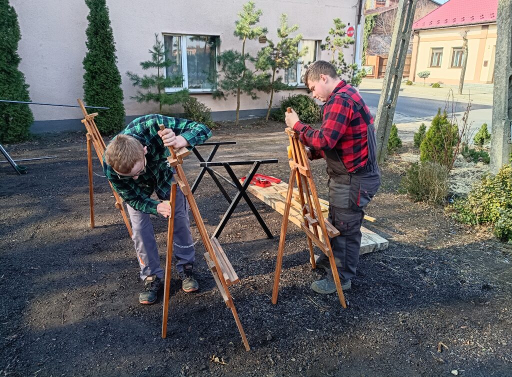 Dwaj młodzi mężczyźni ustawiają drewniane sztalugi na zewnątrz, obok stołu z narzędziami stolarskimi.