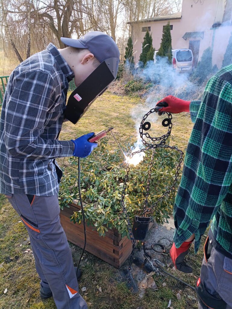Osoba w masce spawalniczej i rękawicach spawa metalową postać stworzoną z łańcuchów, podczas gdy druga osoba trzyma tę postać.