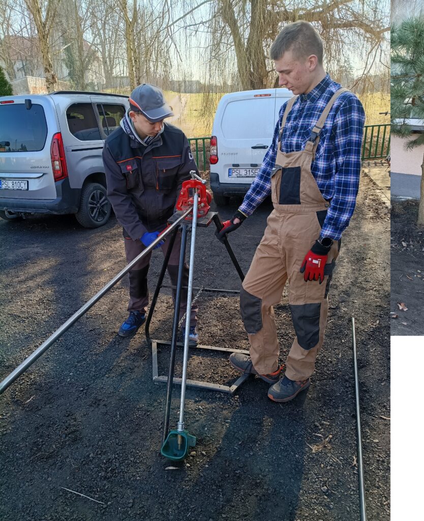 Dwóch mężczyzn w odzieży roboczej pracuje na zewnątrz przy montażu metalowego sprzętu narzędziowego na czarnym podłożu.