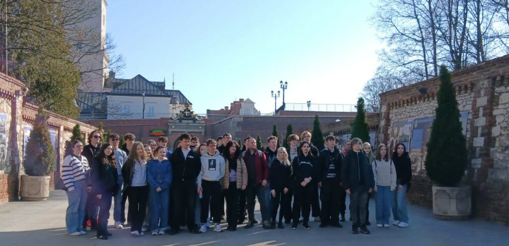 Grupa młodych ludzi stoi razem na tle zabytkowego muru z dekoracyjnymi roślinami w dużych donicach.