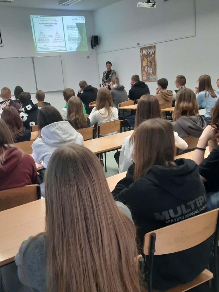 Uczniowie siedzą w ławkach i słuchają nauczycielki omawiającej zmiany w piramidzie żywieniowej wyświetlane na tablicy.
