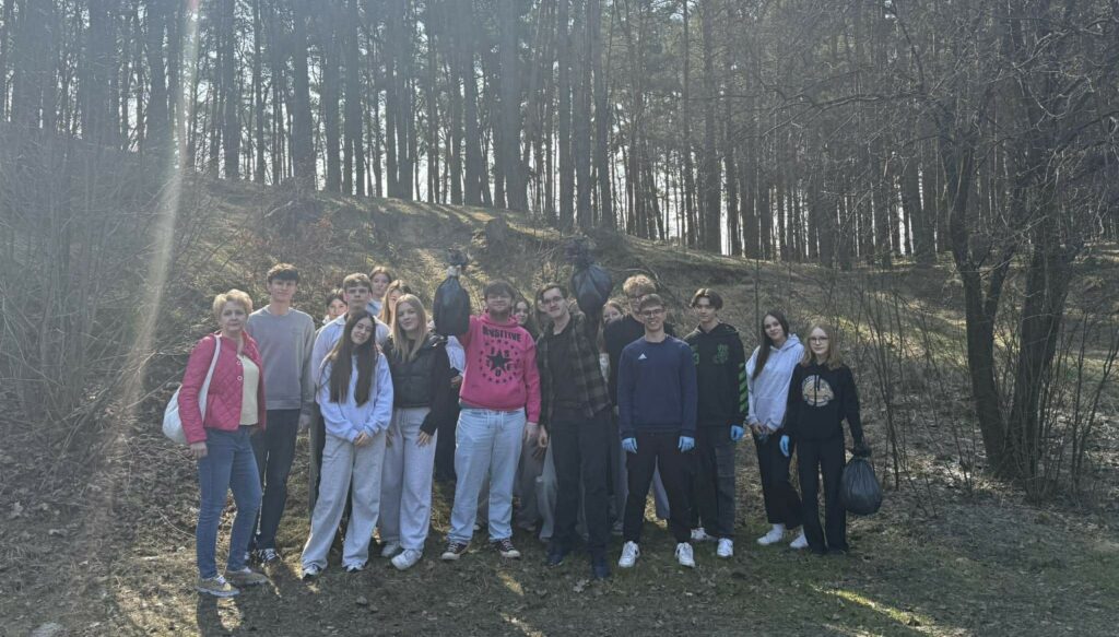 Grupa młodych ludzi i dorosła osoba na tle lasu po sprzątaniu, trzymająca w rękach pełne worki na śmieci.