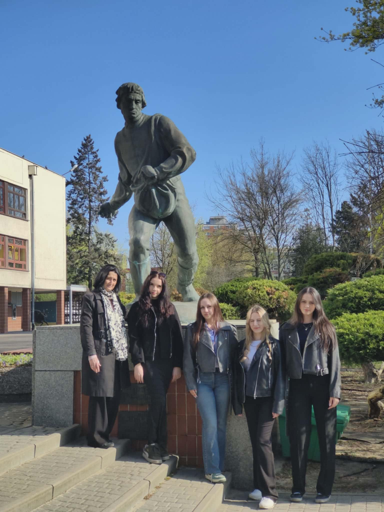 Grupa kobiet stoi przed dużym pomnikiem mężczyzny w dynamicznej pozie na tle budynku i zieleni.