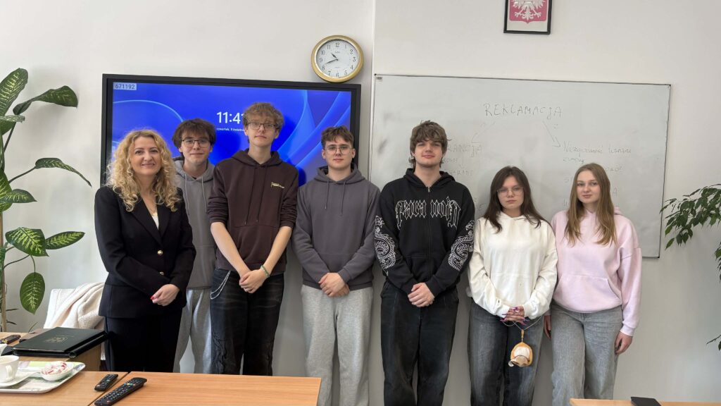 Nauczycielka i sześciu uczniów stoi w klasie przed tablicą z napisem „REKLAMACJA” oraz monitorem.