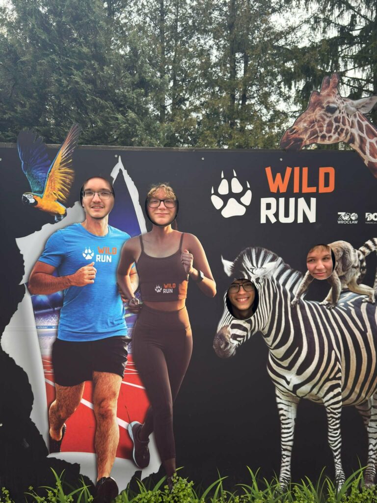 Cztery osoby biorą udział w zabawie z tablicą WILD RUN, gdzie ich twarze wstawione są w sylwetki biegaczy i zwierząt – zebry i lemura.