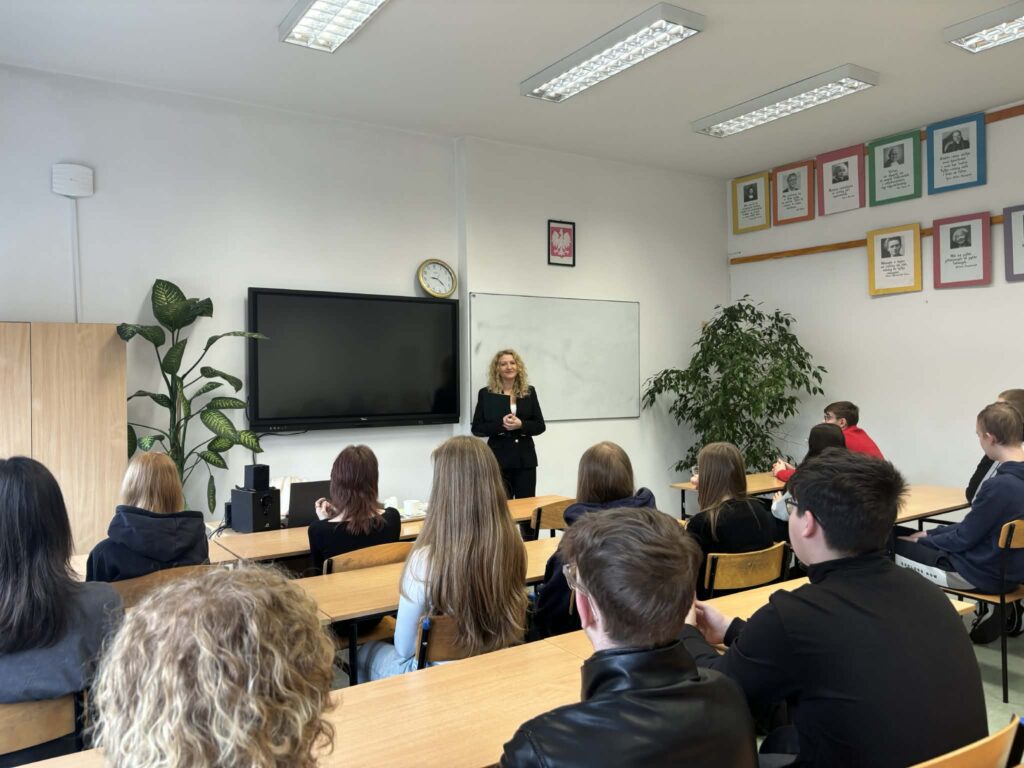 Nauczycielka przemawia do uczniów siedzących przy ławkach w klasie szkolnej.