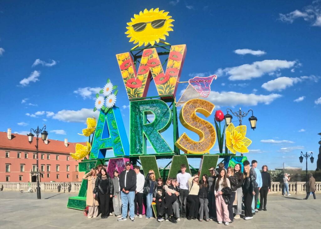 Kolorowa instalacja z napisem "WARSZAWA" ozdobiona kwiatami i uśmiechniętym słońcem w okularkach, a przed nią stoi grupa młodych ludzi na tle jasnego, słonecznego nieba.