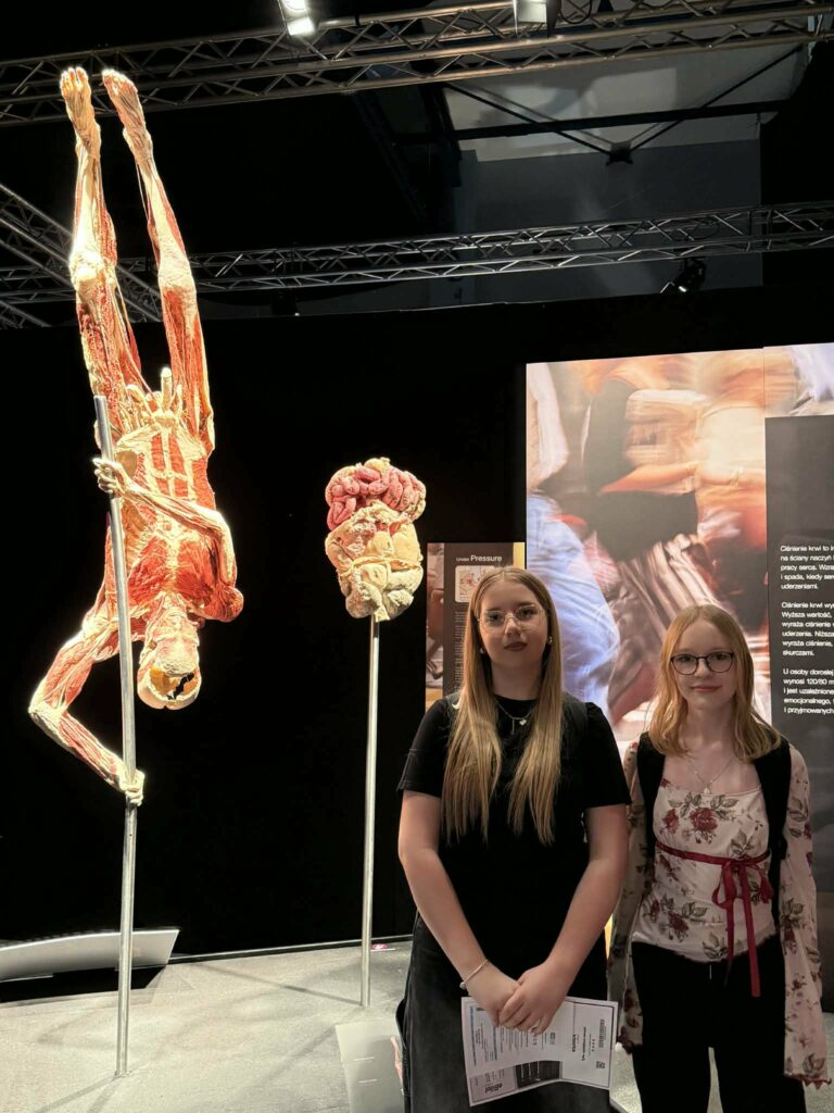 Dwie młode kobiety stoją w muzeum obok eksponatów anatomicznych, w tym wiszącej odwróconej postaci ludzkiej bez skóry.