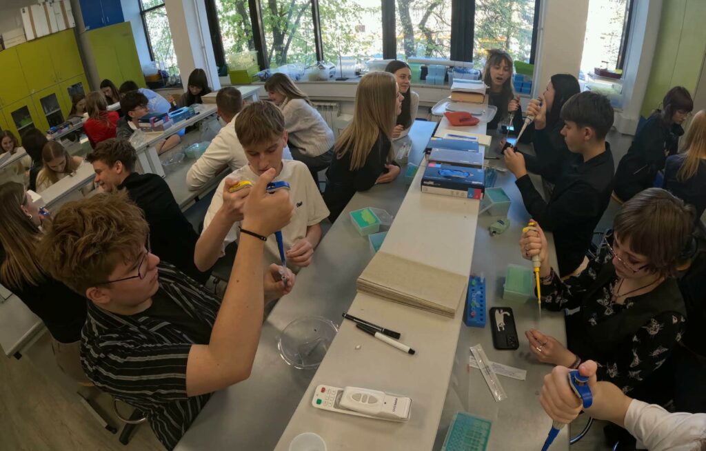 Grupa uczniów w pracowni laboratoryjnej wykonuje eksperymenty z użyciem pipet i probówek.