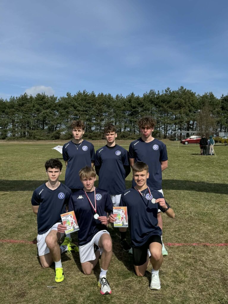 Sześciu młodych sportowców w granatowych strojach drużynowych na otwartym boisku, trzech z nich trzyma pamiątkowe vouchery i medale.