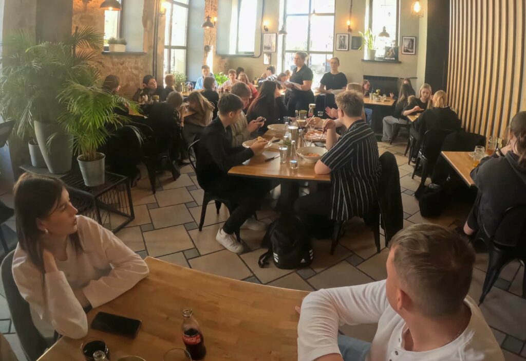 Wnętrze zatłoczonej restauracji z młodymi ludźmi jedzącymi i rozmawiającymi przy drewnianych stołach oraz obsługą w ruchu.