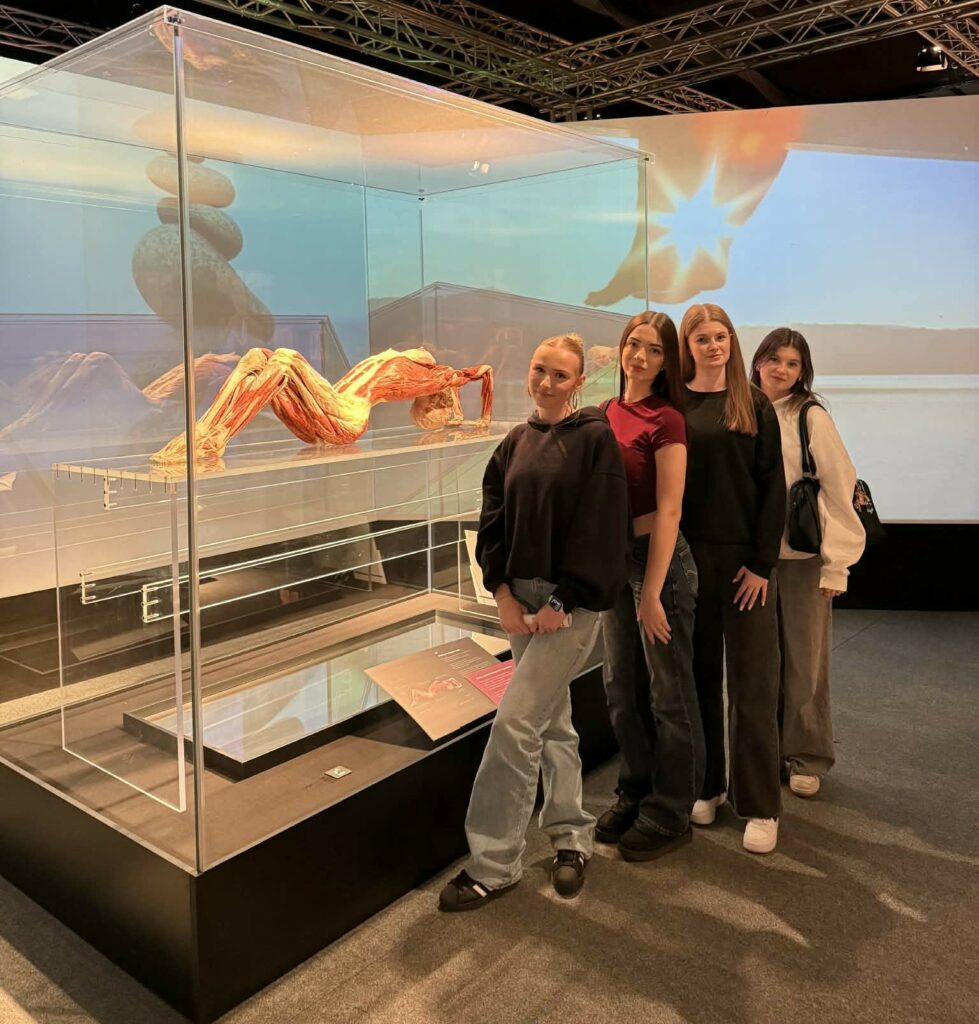 Cztery młode kobiety stoją obok oszklonej ekspozycji z modelem anatomicznym ludzkiego ciała, na tle projekcji natury ze słońcem.