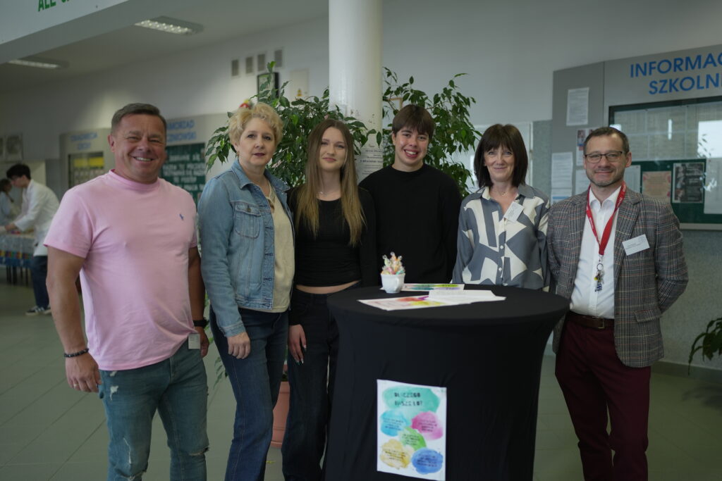 Grupa sześciu osób stoi razem przy okrągłym stole z ulotkami w szkolnym korytarzu.