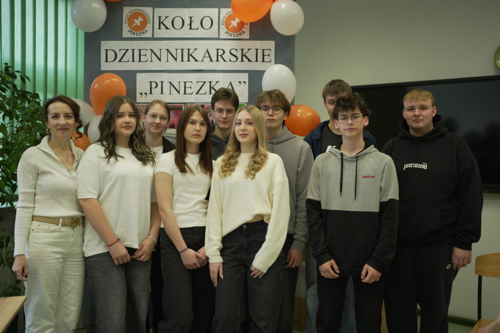 Grupa młodzieży i nauczycielka stoją razem na tle dekoracji z balonami i napisem „Koło Dziennikarskie Pinezka”.