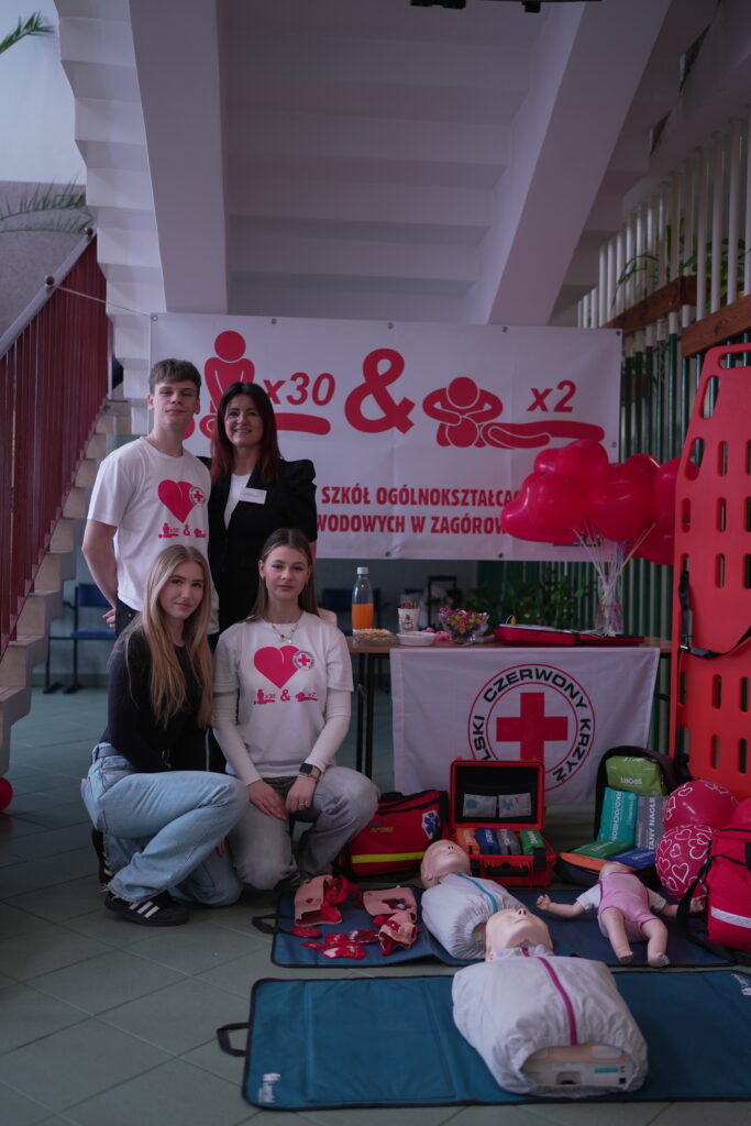 Grupa młodych osób na stoisku Polskiego Czerwonego Krzyża z manekinami i materiałami do nauki pierwszej pomocy.